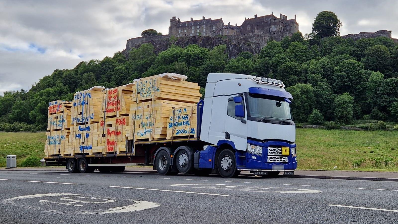 White and blue articulated lorry loaded with unwrapped timber frame kit packs marked with plot numbers, parked on a road below Stirling Castle