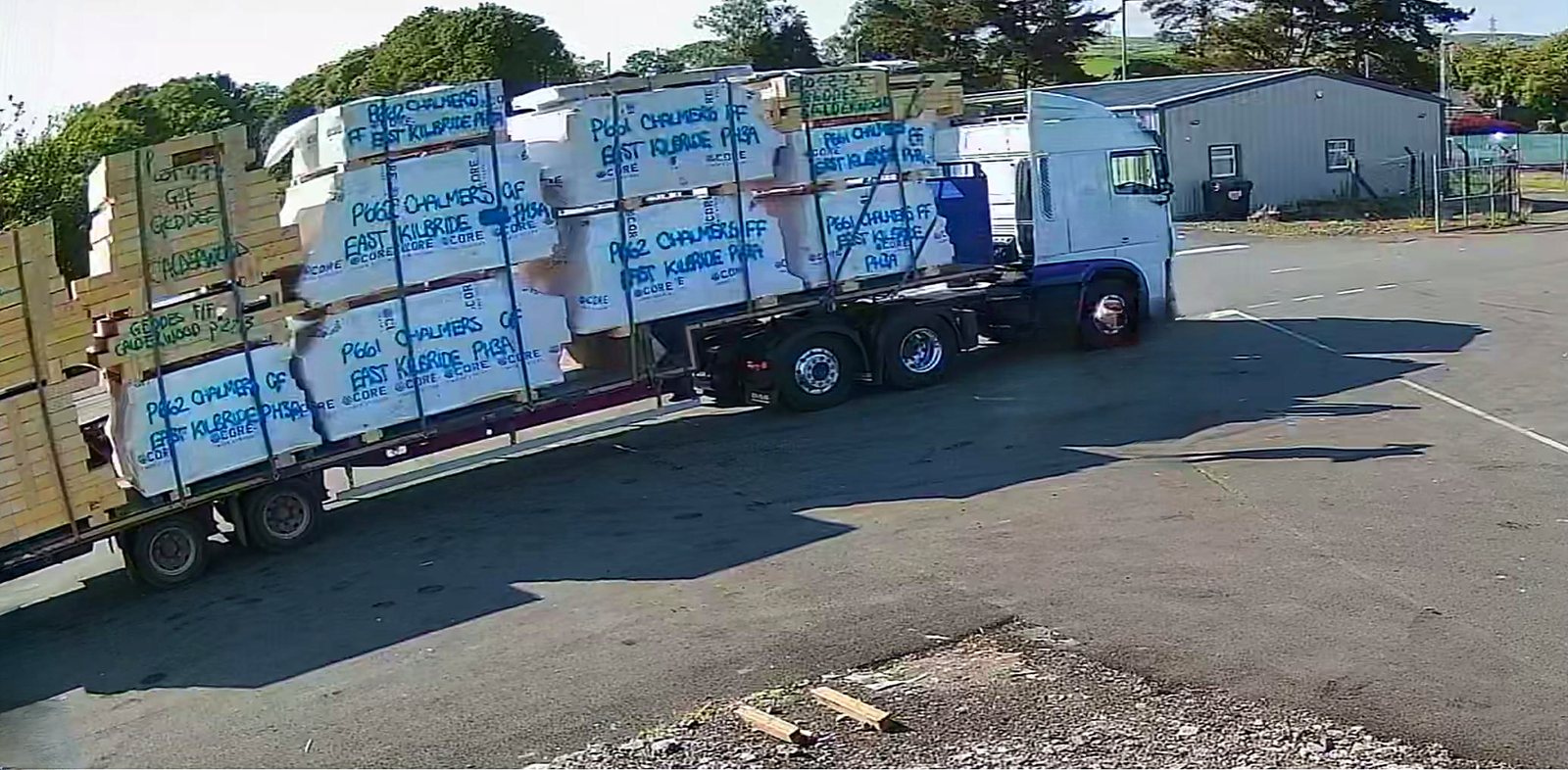 Articulated lorry in a factory yard loaded with timber frame kit packs wrapped in white Core-branded wrap, with plot details handwritten in blue on each pack