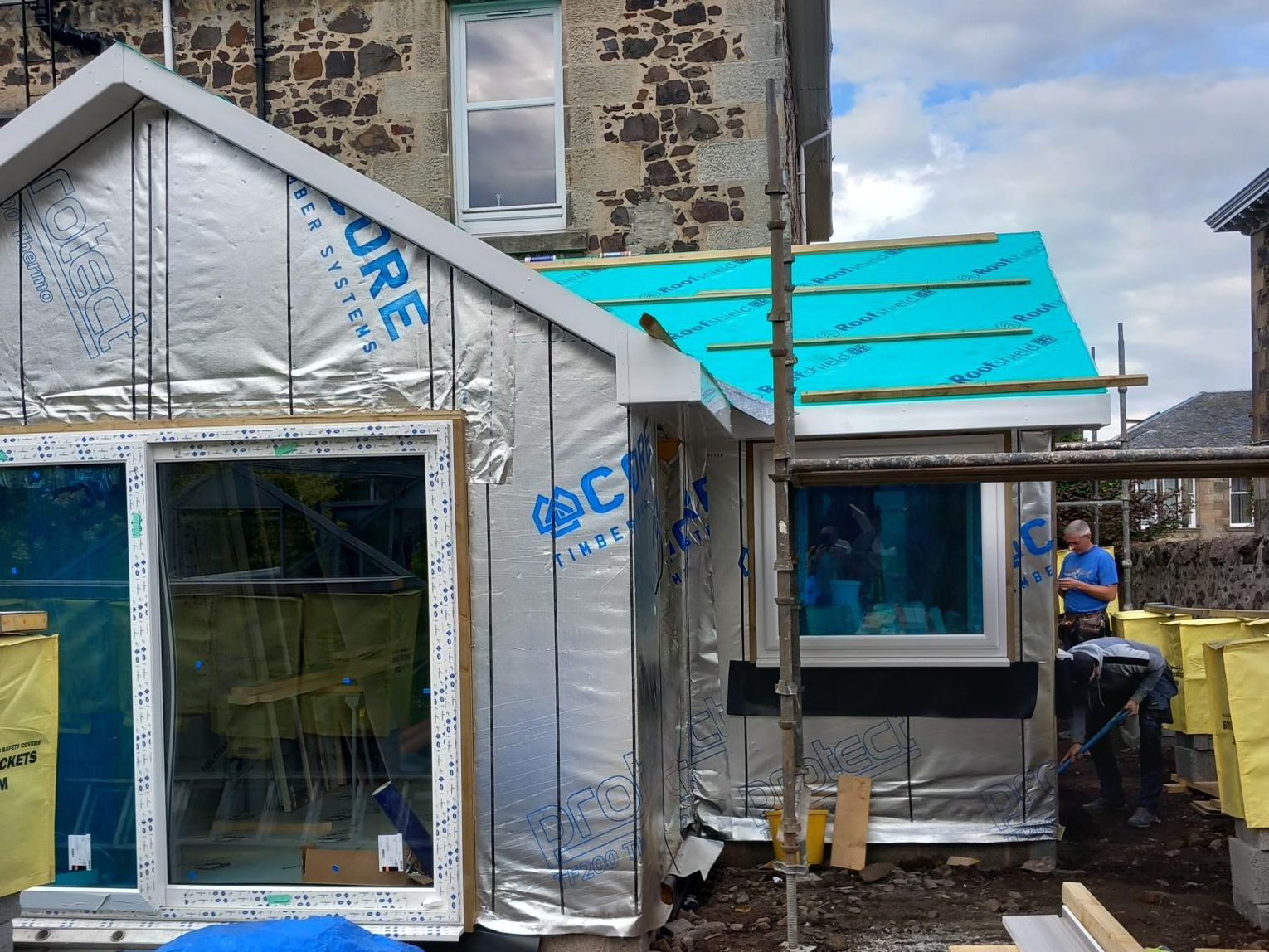 Timber frame extension under construction at the back of a traditional stone house, wrapped in breather membrane printed with Core Timber Systems branding, with a large window frame fitted in the gable, roofing underlay on the lean-to section, workers on site and timber stacked in the foreground