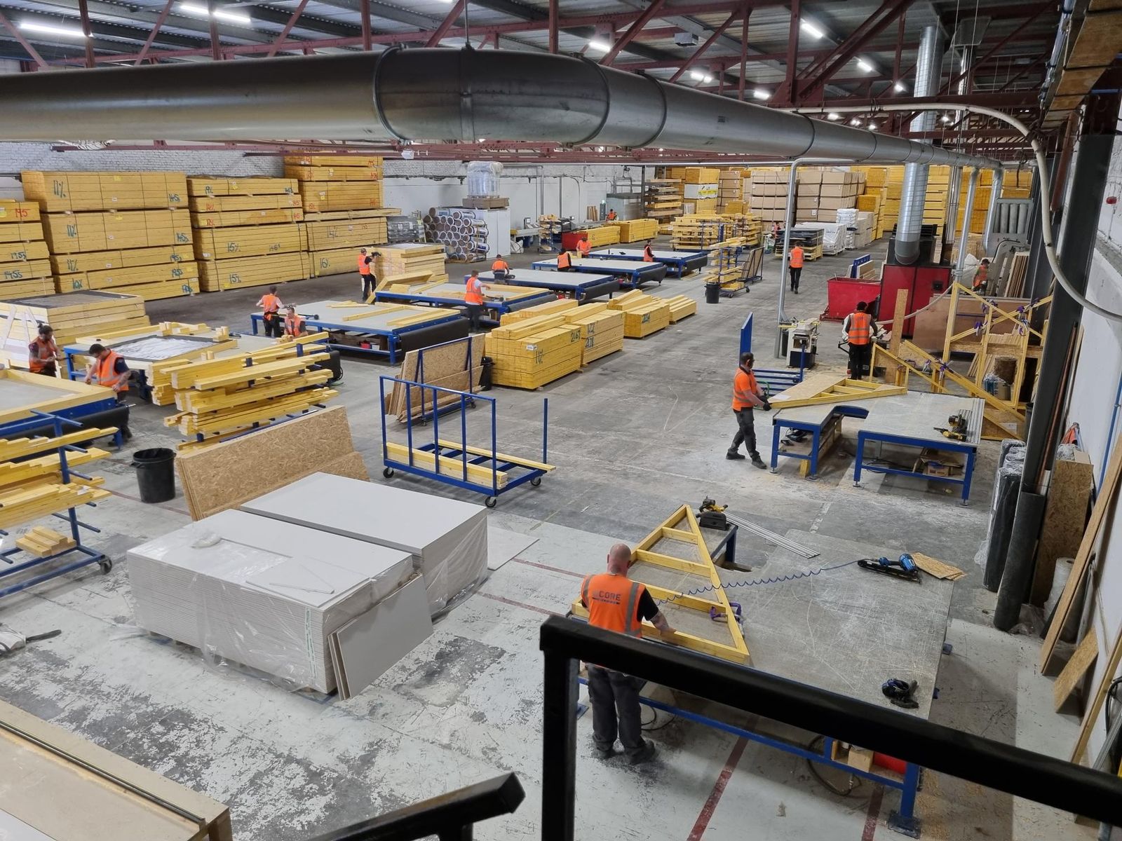 Factory floor seen from a mezzanine: staff in orange hi-vis building timber frame wall panels and roof trusses on blue workbenches, with stacks of wrapped timber packs, OSB and plasterboard sheets, and extraction ducting overhead