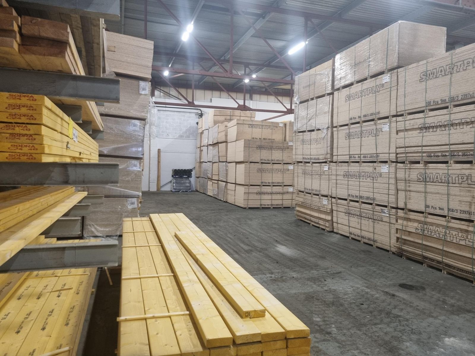 Timber store inside the factory: cantilever racks of graded structural timber on the left, a pack of timber in the foreground and tall stacks of pallets of OSB sheathing board on the right