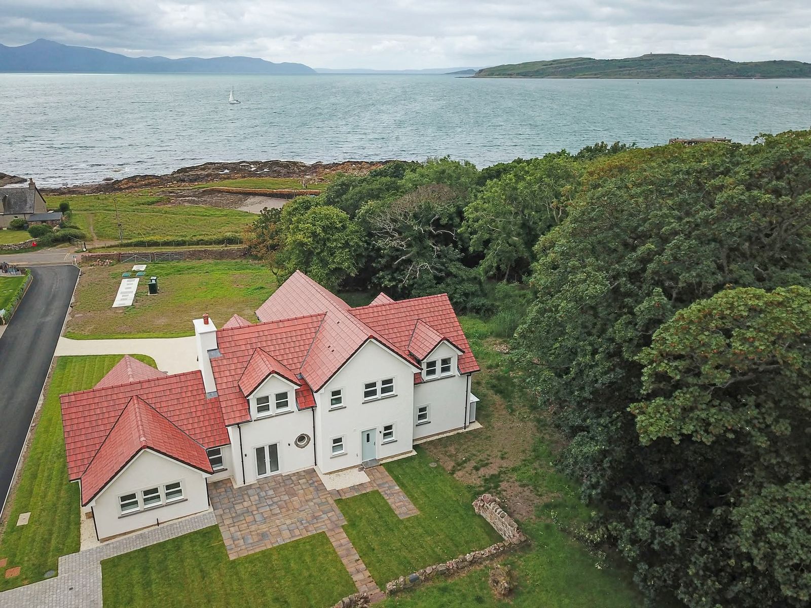 Aerial view of a finished white-rendered house with red tiled roofs in Portencross, North Ayrshire, set among trees beside the sea with hills and an island behind