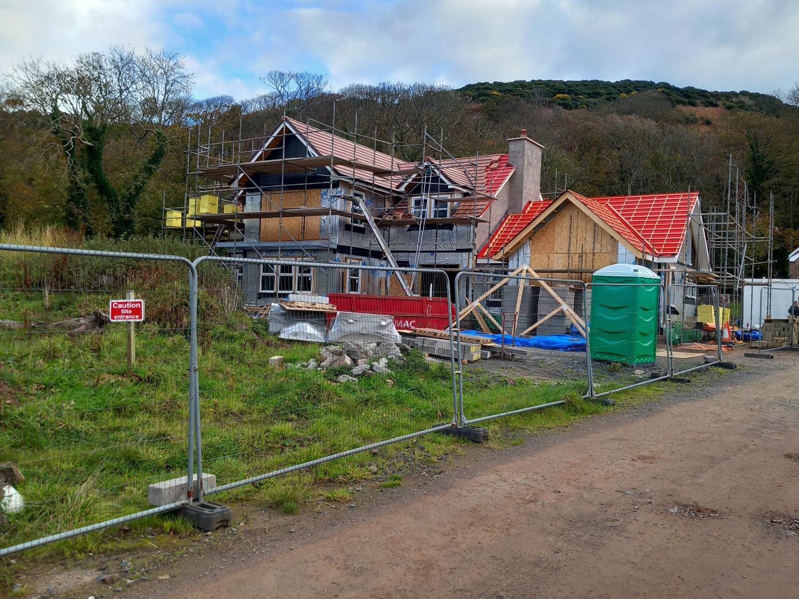 The same house during construction: the timber frame up, sheathed and wrapped in membrane, with full scaffolding, red roof tiles going on, site fencing, a skip and a portable toilet in the foreground