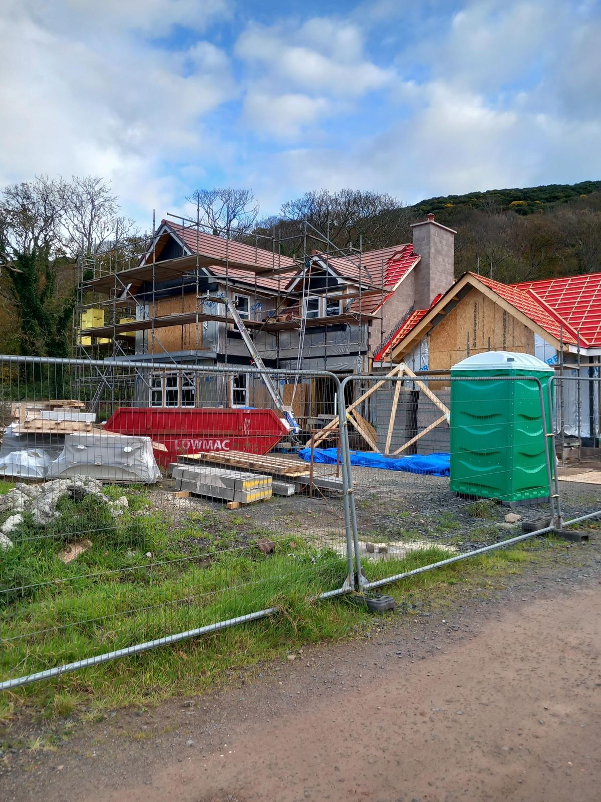 Closer view of the house during construction, with the scaffolded timber frame, roof tiles being laid on the single-storey wing and spare roof trusses on site behind the fencing