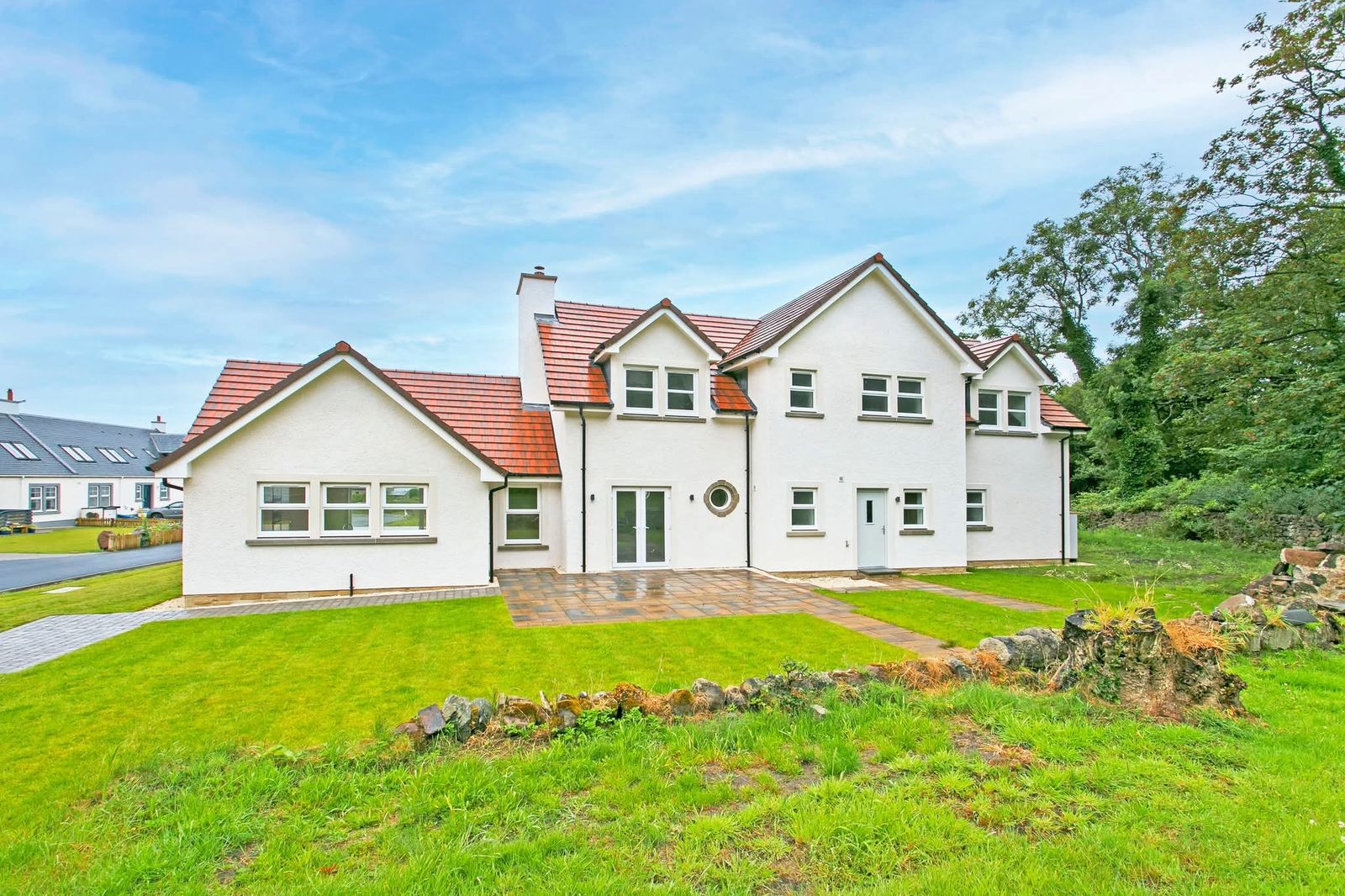 Finished detached house with white render, red tiled roofs, several gables, a round porthole window, a paved patio and lawn