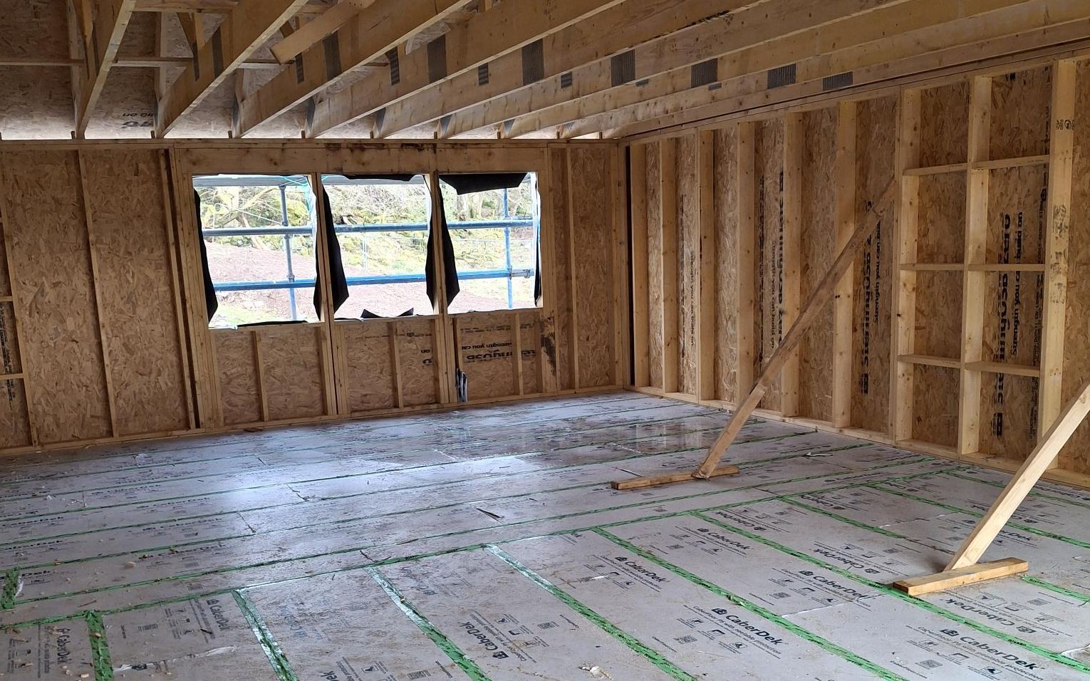 Inside a timber frame house under construction: OSB-sheathed wall panels with window openings, metal-web floor joists overhead, and the floor deck laid with taped joints and temporary bracing