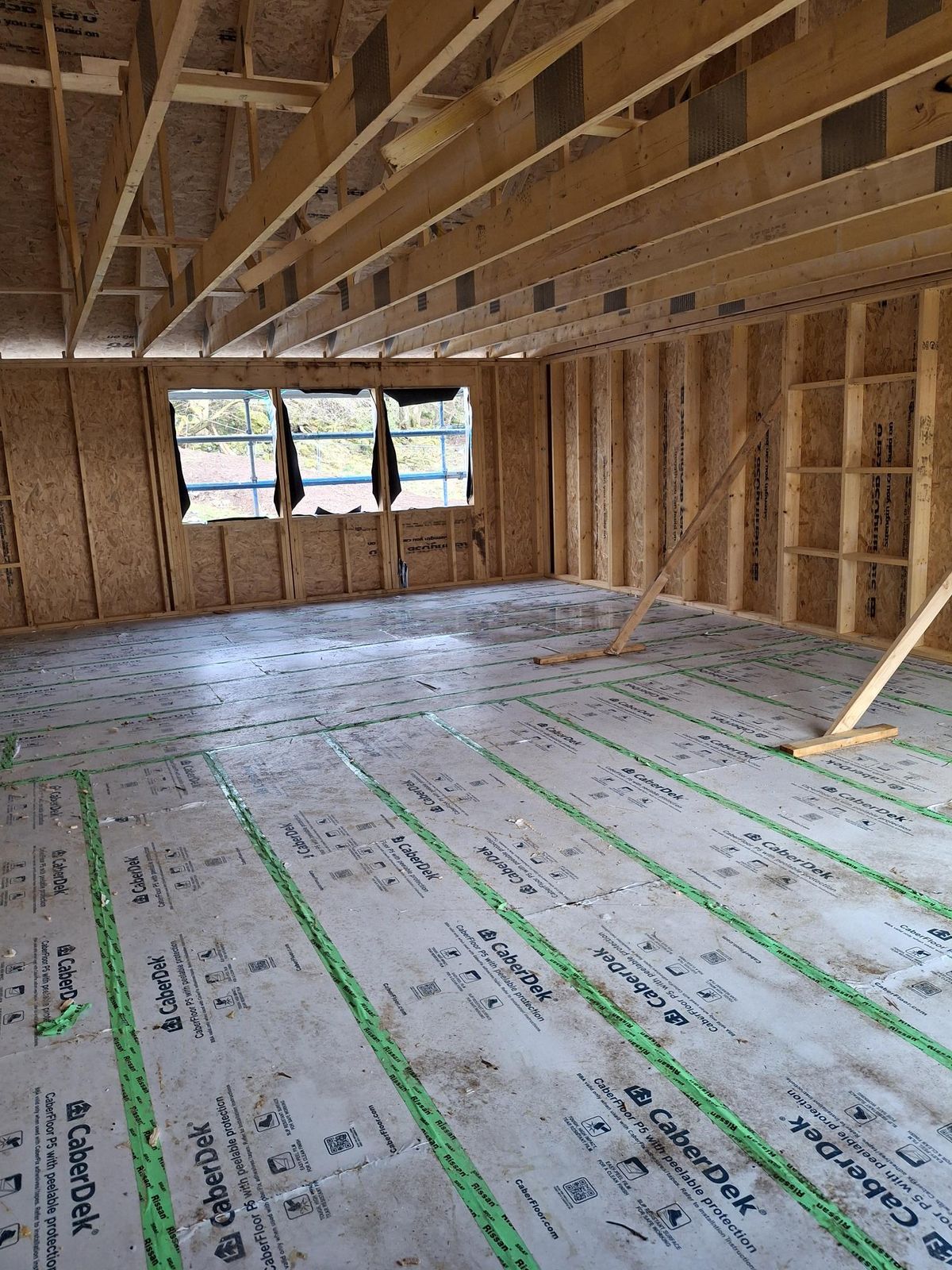 Inside a timber frame house under construction: OSB-sheathed wall panels with window openings, metal-web floor joists overhead, and the floor deck laid with taped joints and temporary bracing