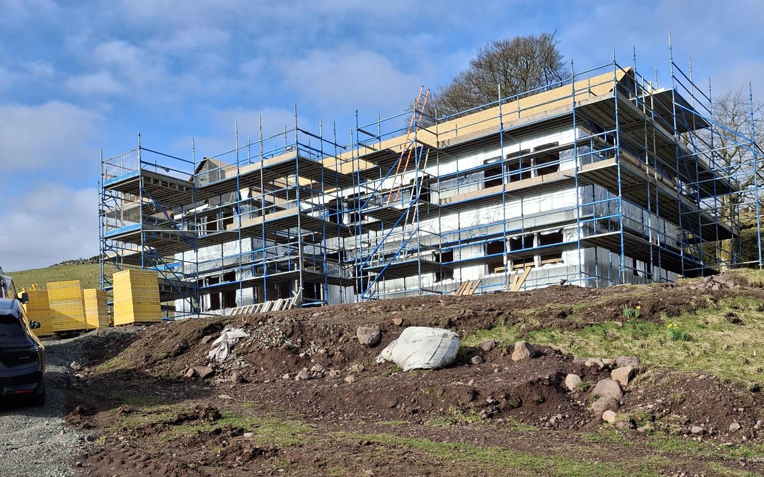 Large timber frame building under construction on a hillside site, fully scaffolded with the frame and sheathing up and membrane on, timber packs alongside and a gravel track in the foreground