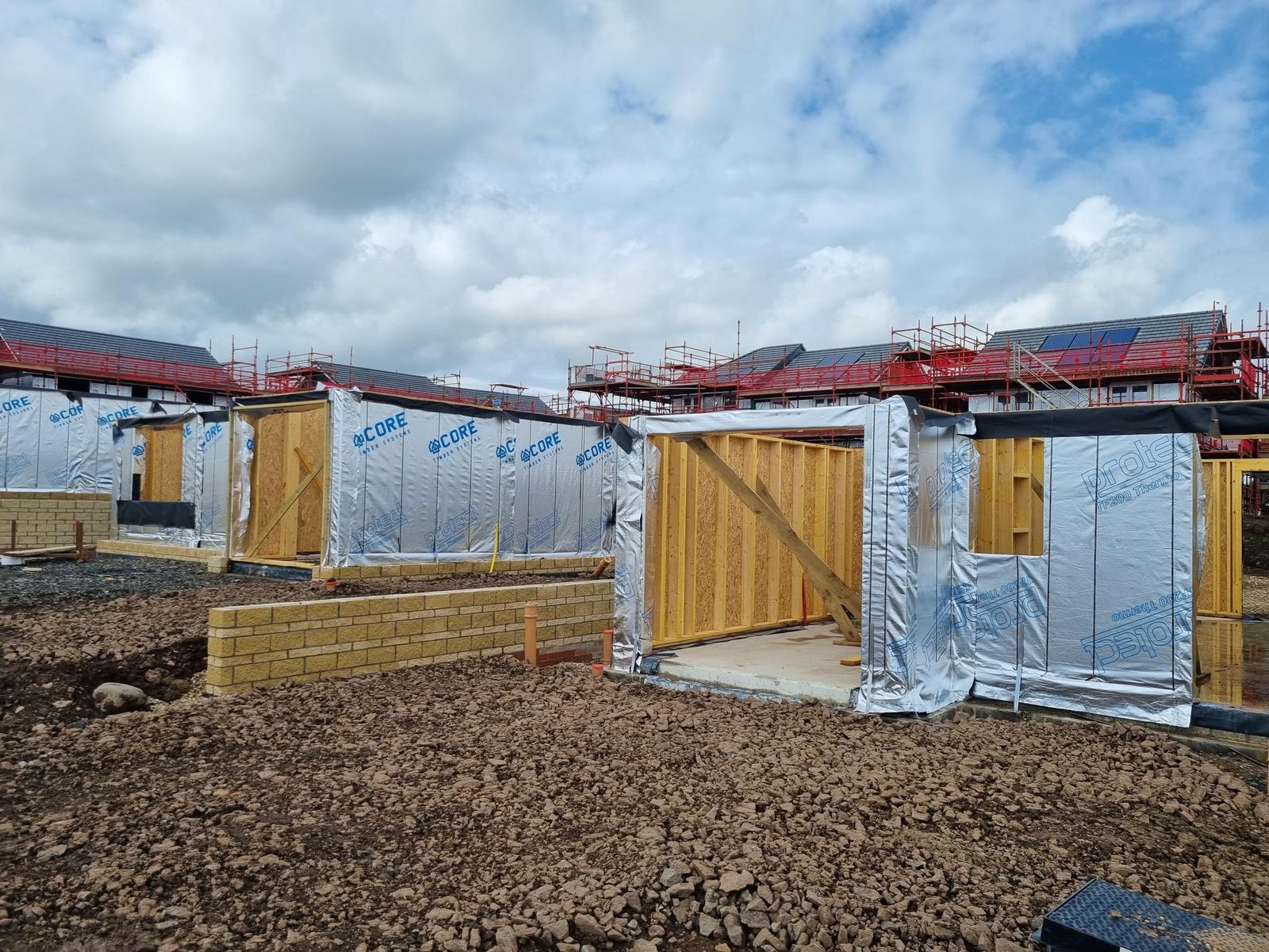 Ground floor open panel timber frame wall panels erected on concrete slabs on a Taylor Wimpey housing development, wrapped in breather membrane printed with the Core Timber Systems logo, with blockwork walls in front and scaffolded houses behind