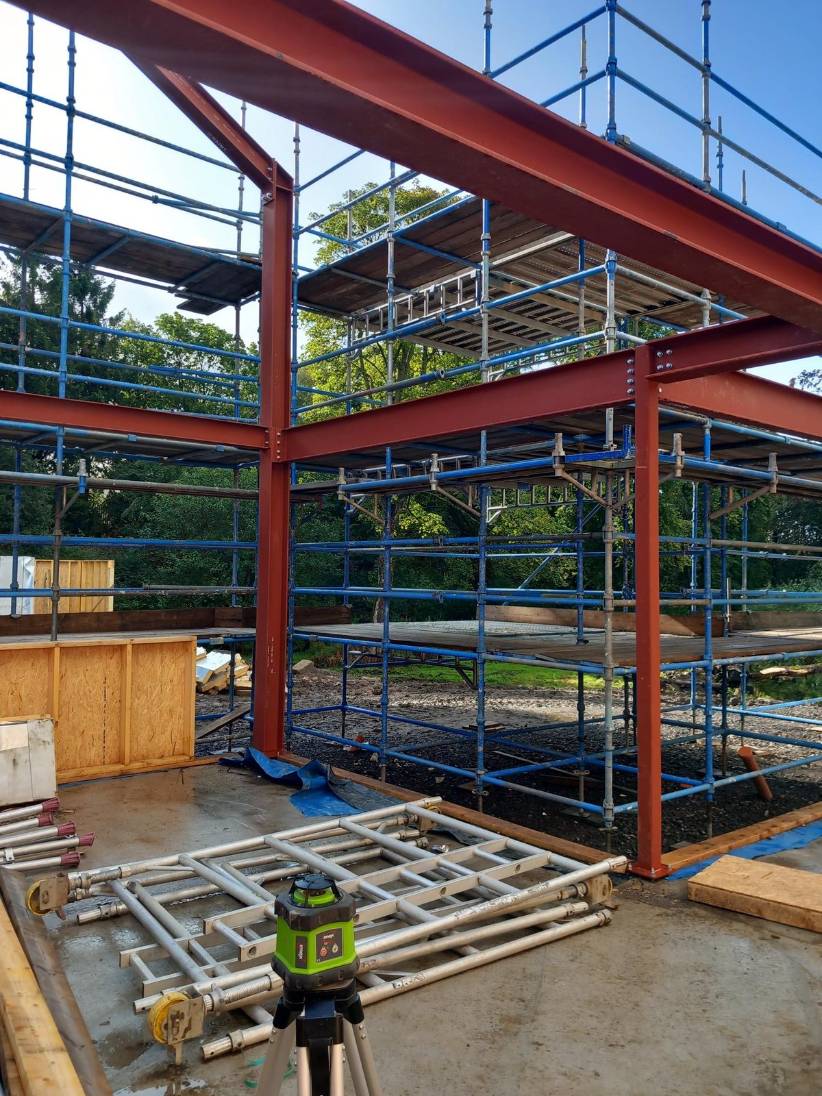 Red structural steel frame on a concrete slab, surrounded by blue scaffolding, with a laser level on a tripod in the foreground