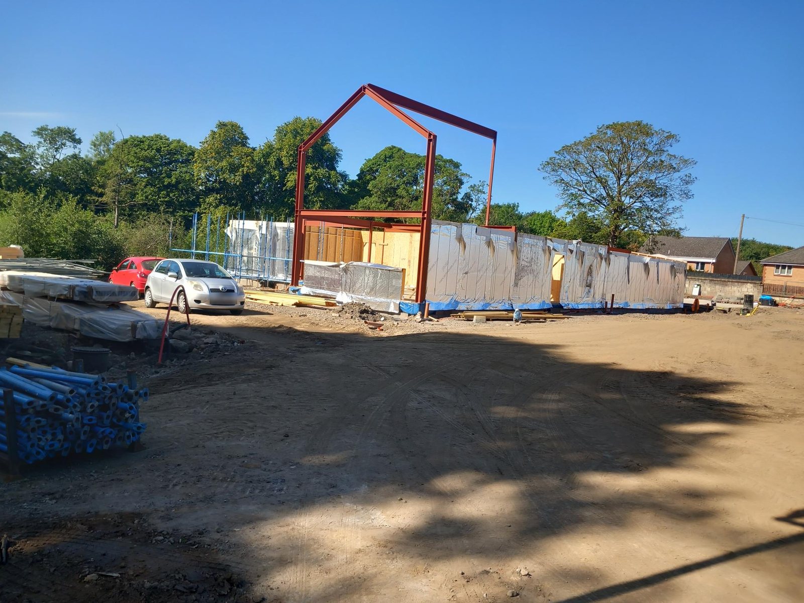 Wide view across the Newmains self-build site: a red steel portal frame above timber frame walls wrapped in breather membrane, with trees behind and a gravel site in the foreground