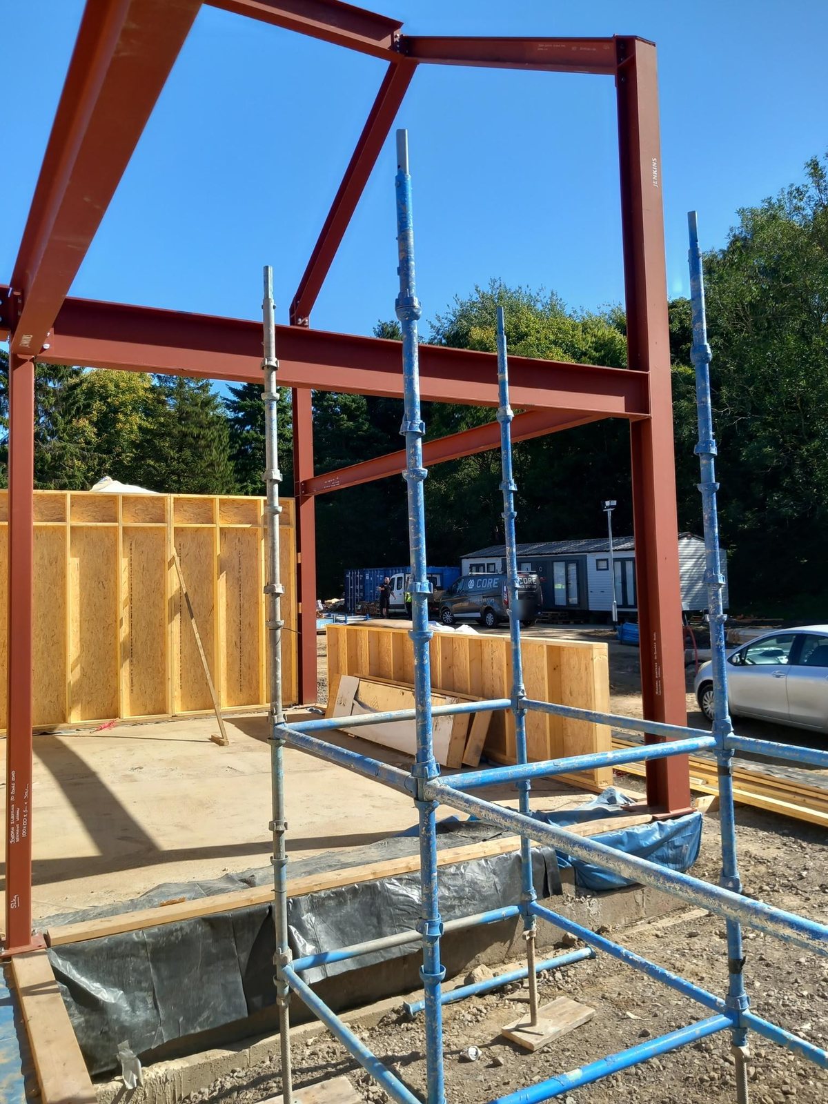 Red steel frame and scaffolding with the first OSB-sheathed open panel wall panels standing on the slab, and a Core Timber Systems branded van in the background