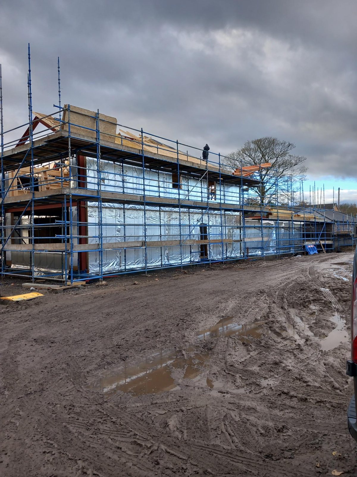Two-storey timber frame wrapped in foil breather membrane and fully scaffolded, with roof timbers going on, seen across a muddy site