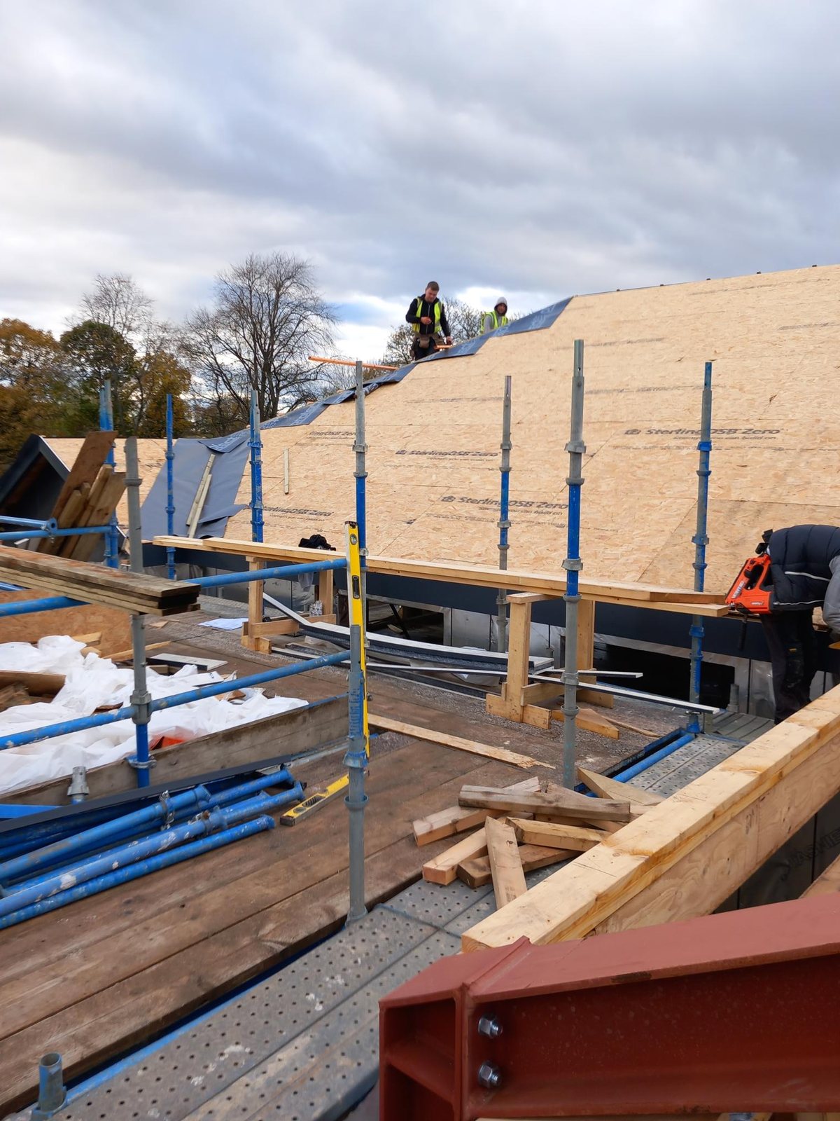 Roof sheathed in OSB with two workers in hi-vis on it, seen from the scaffold with timber and a red steel beam in the foreground