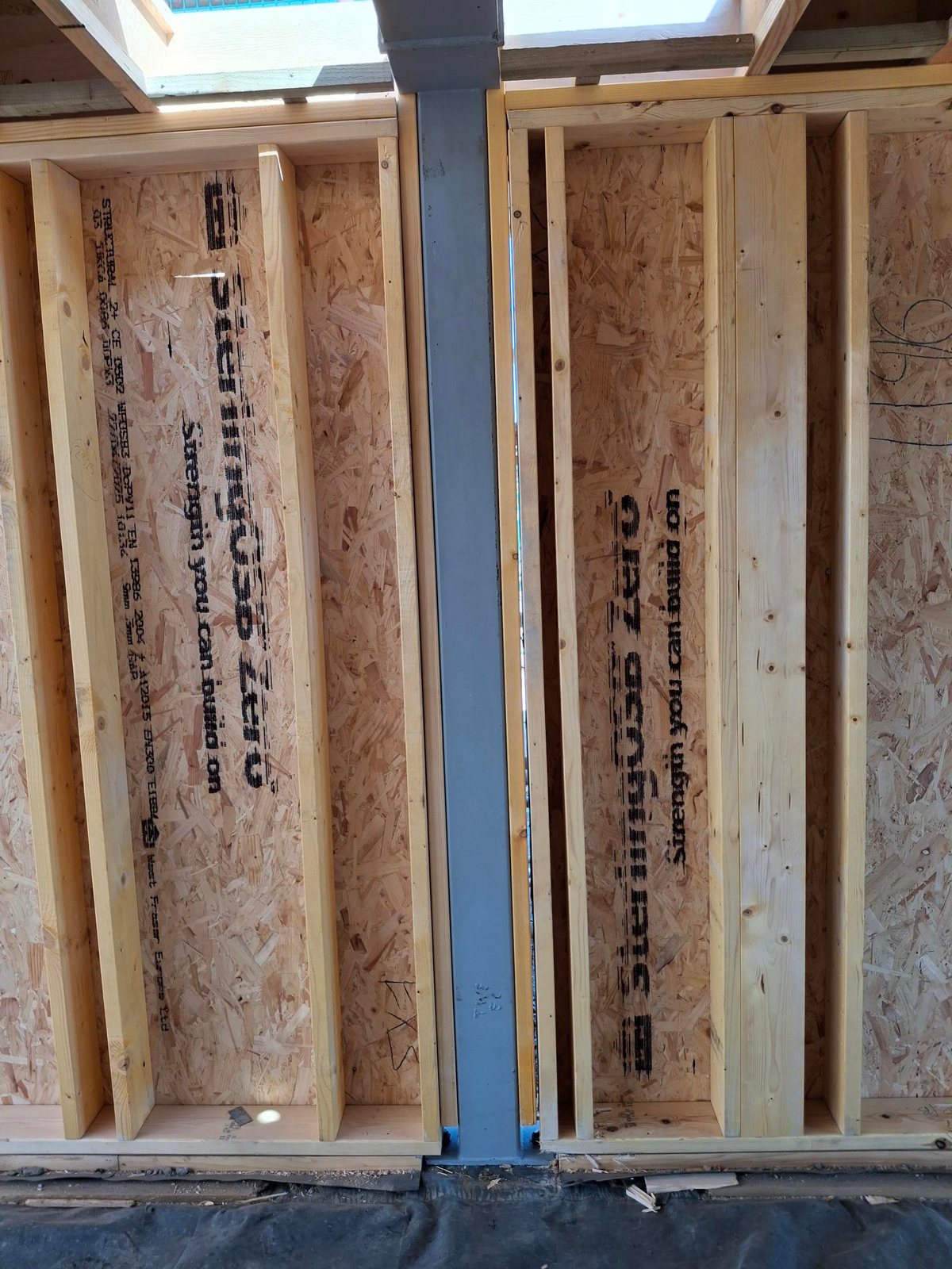 Close-up of open panel timber frame wall studs with OSB sheathing on either side of a grey steel post