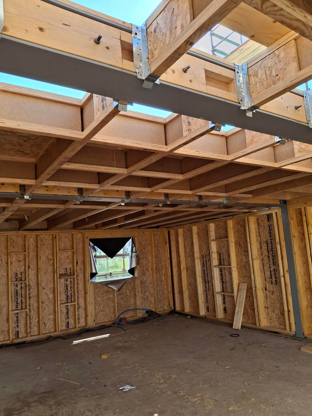 Ground floor of a timber frame house under construction: metal-web floor joists overhead fixed to a steel beam with metal joist hangers, and OSB-sheathed open panel stud walls around the room